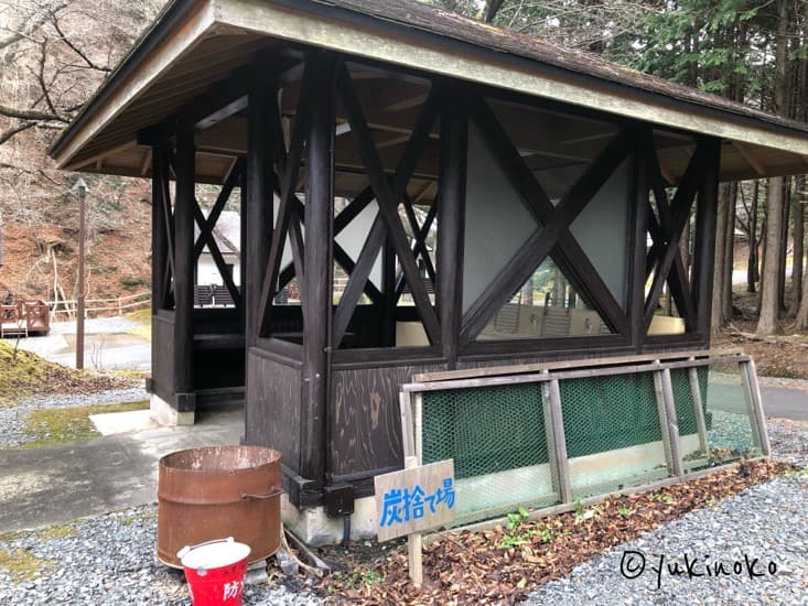 木でできた屋根の小屋のような見た目の炊事場、手前にはドラム缶とバケツが置いてあり、炭捨て場と書いた看板が隣にある