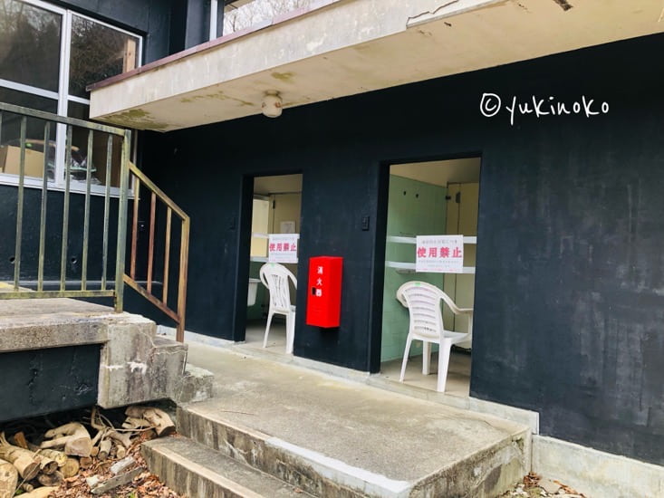 黒い壁の平屋のトイレの建物にドア無の入り口が二つあり、入り口に白い椅子がそれぞれ1脚ずつ置かれている