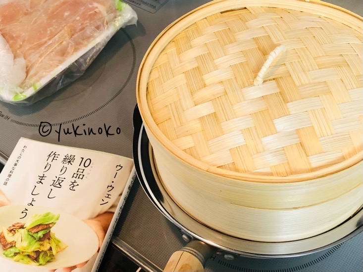 片手鍋の上に蒸し板とせいろが置かれていて、奥にはトレイに入った鶏肉と、手前には本が置いてある