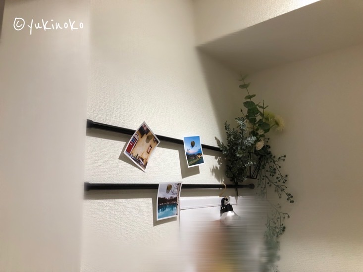 白い壁に2本の黒い突っ張り棒が横に付けられていて、突っ張り棒には写真が3枚磁石で貼られていて、右端には造花や、真ん中あたりにはS字フックでカレンダーがぶら下げられている。
