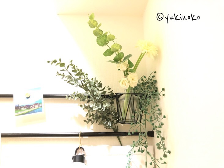 つっぱり棒に、手のひらサイズの目が荒いカゴが引っ掛けてあり、カゴには小瓶が入れられ、造花が飾られている