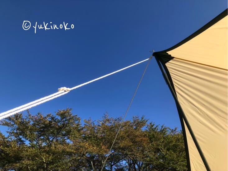 タープを張った状態を下から見上げていて、青空を背景にタープの先端部分から紐が繋がって伸びている様子が見える