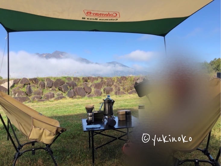 青空と山を背景に、ポール2本とロープを使用してタープが張られている様子、タープの下にテーブルと椅子が置いてある