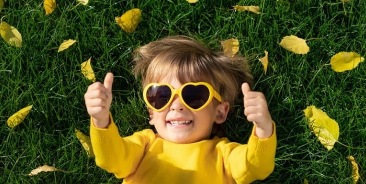 芝生に黄色い服とフチが黄色いサングラスをかけた子どもが寝っ転がり、上半身だけ中央に写っていて、こちらを向いて両手でグッドサインをしながら笑っている。芝生の上には黄色い落ち葉がいくつか落ちている。