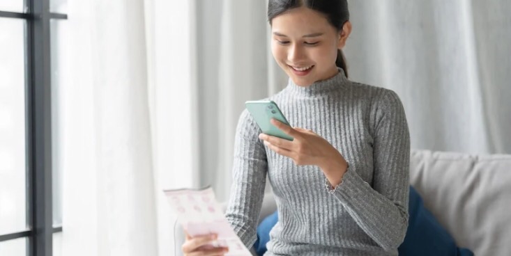 右手にレシート、左手に携帯電話を持って、携帯を見ながら笑顔になっている女性がソファーに座っている