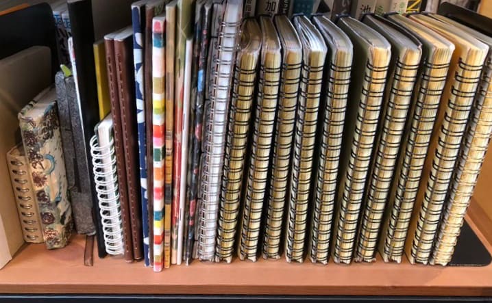 本棚にノートが置いてあり、左側はサイズの異なるノートが数十冊、右側に行くとリング式の同じノートが数十冊ほど並んでいる